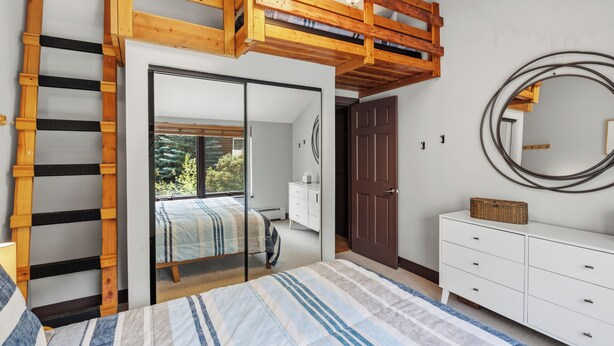 Un dormitorio con cama doble alta, un armario, un espejo y vistas a través de un ventanal.