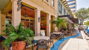 An outdoor cafe with tables and chairs, a large potted plant, and a building with a sign above the entrance.