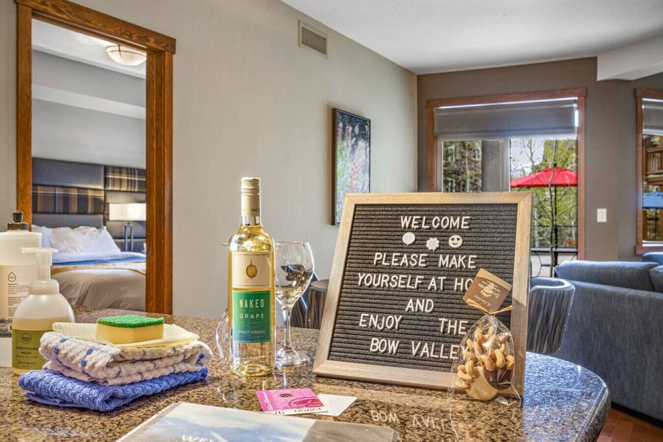 Habitación de hotel con una cama, una botella de vino Naked Grape, una copa, toallas, un cartel de bienvenida y vistas al exterior.