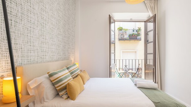 A neatly arranged bedroom with a bed, bedside lamp, and a view of a balcony with plants and a chair. - Apartamentos Luna Suites Palacio Dorita (Granada)