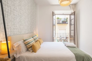 Ein ordentlich eingerichtetes Schlafzimmer mit Bett, Nachttischlampe und Blick auf einen Balkon mit Pflanzen und einem Stuhl.