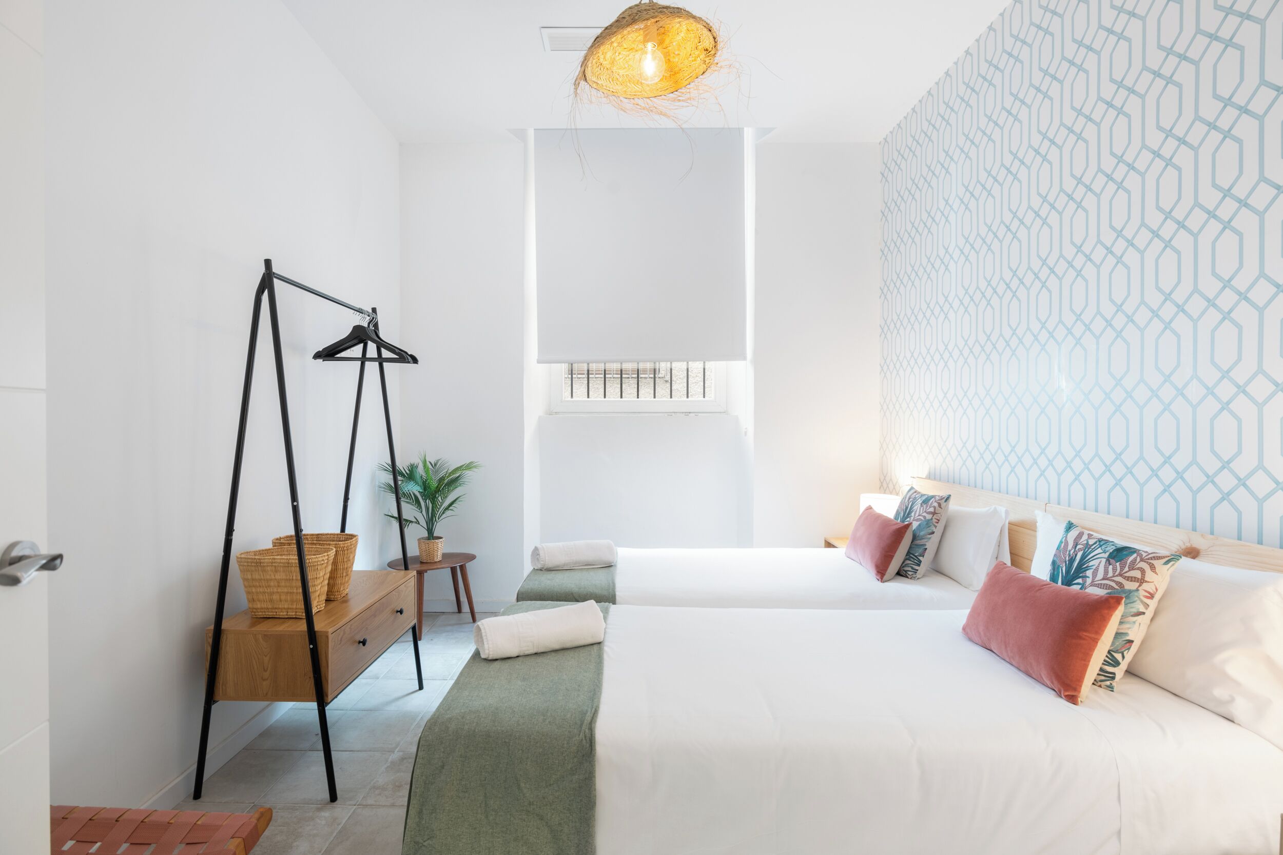 A hotel room with a bed, a hanging light fixture, a wooden bench, a small table with a plant, and a patterned wall.