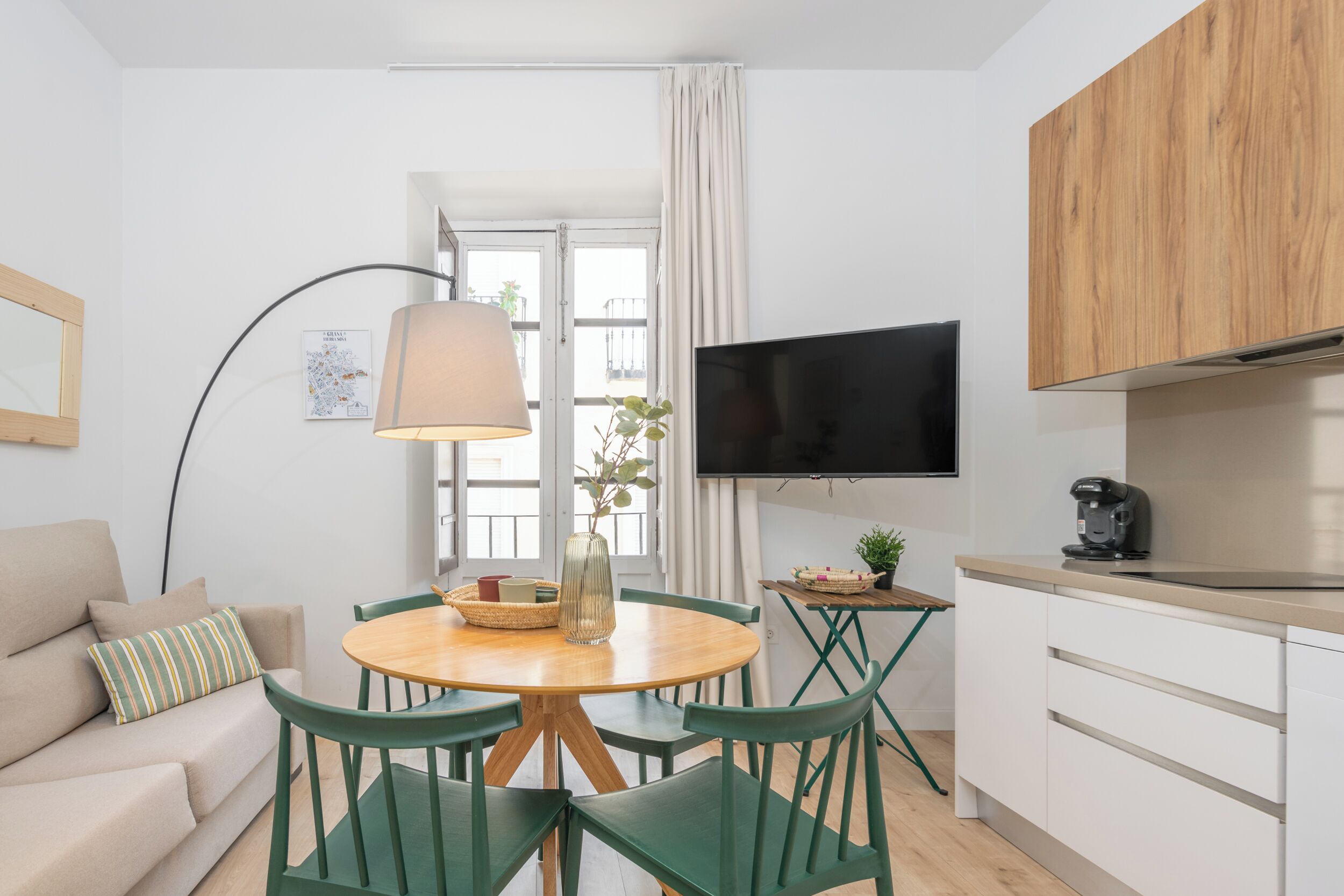Una sala de estar moderna con una mesa redonda de madera, sillas verdes, un sofá beige, una lámpara de pie, un televisor de pantalla plana y una zona de cocina con armarios blancos y una encimera de madera.