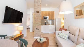 Uma sala de estar moderna com uma televisão de tela plana, uma mesa de centro de madeira, um sofá com almofadas e uma área de cozinha ao fundo.