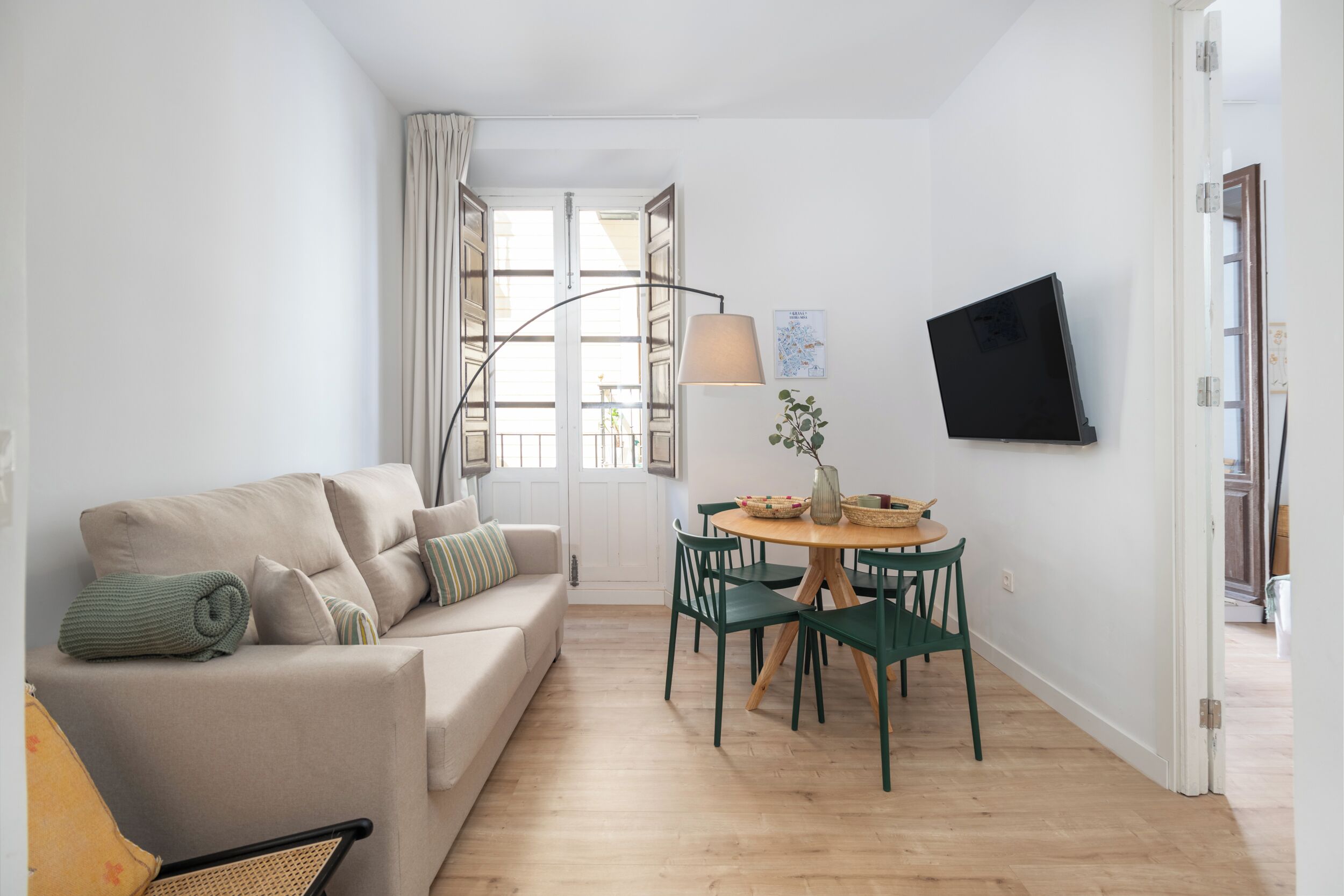A modern living room with a sofa, a dining table with chairs, a lamp, and a TV mounted on the wall.