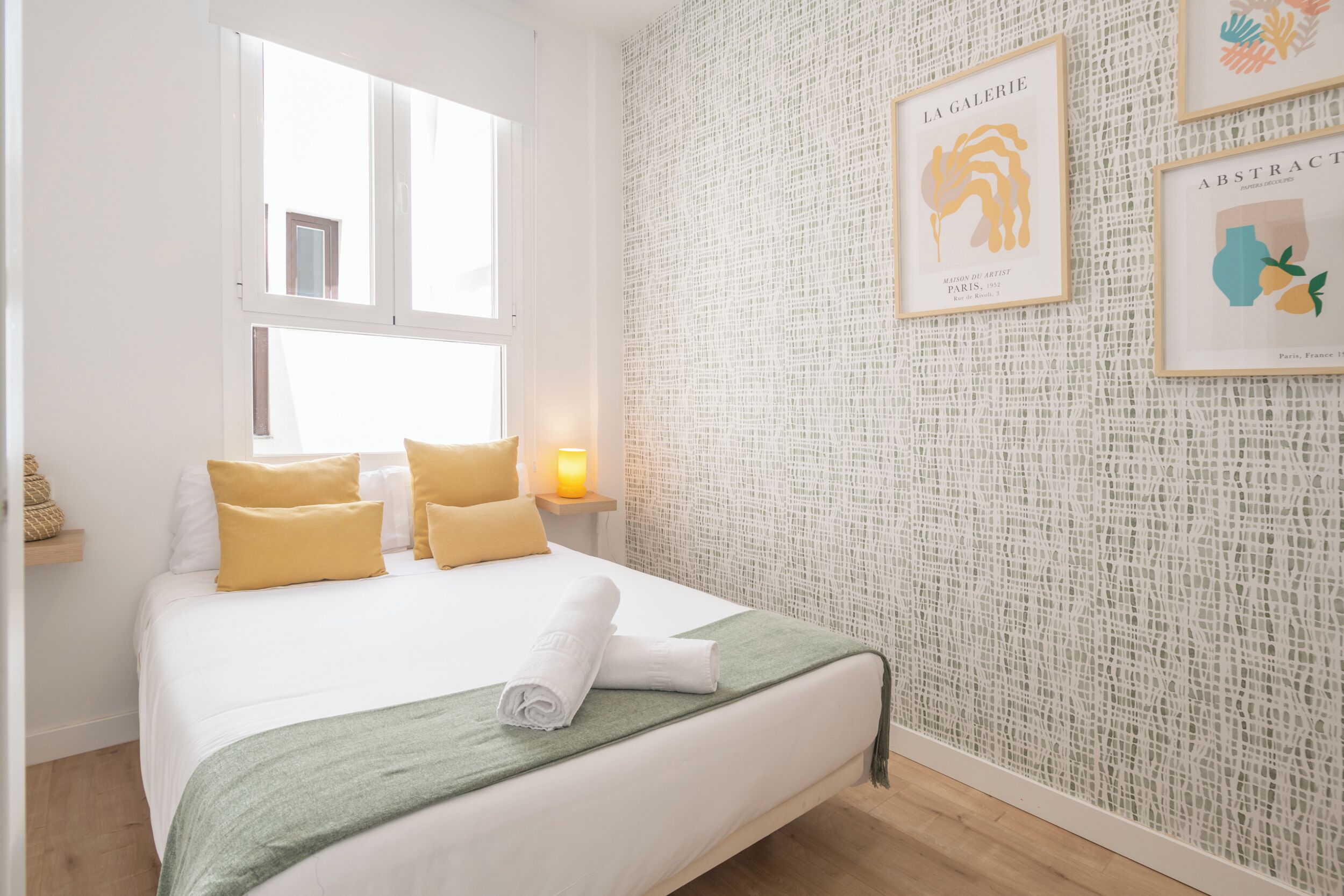 Un lit bien fait, agrémenté d’oreillers jaunes, d’un plaid vert et d’une serviette enroulée. Une fenêtre se trouve au-dessus du lit, et un mur est décoré d’œuvres d’art encadrées.