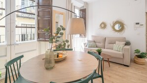 A modern living room with a round wooden table, green chairs, a beige sofa, and a large window with a view of a balcony. - Apartamentos Luna Suites Palacio Dorita (Granada)