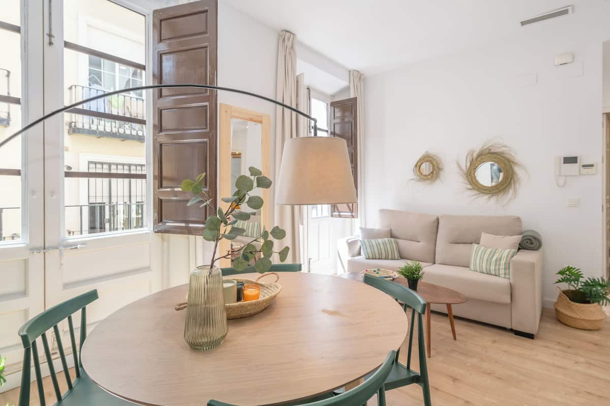 Un salon moderne avec une table ronde en bois, des chaises vertes, un canapé beige et une grande fenêtre donnant sur un balcon.