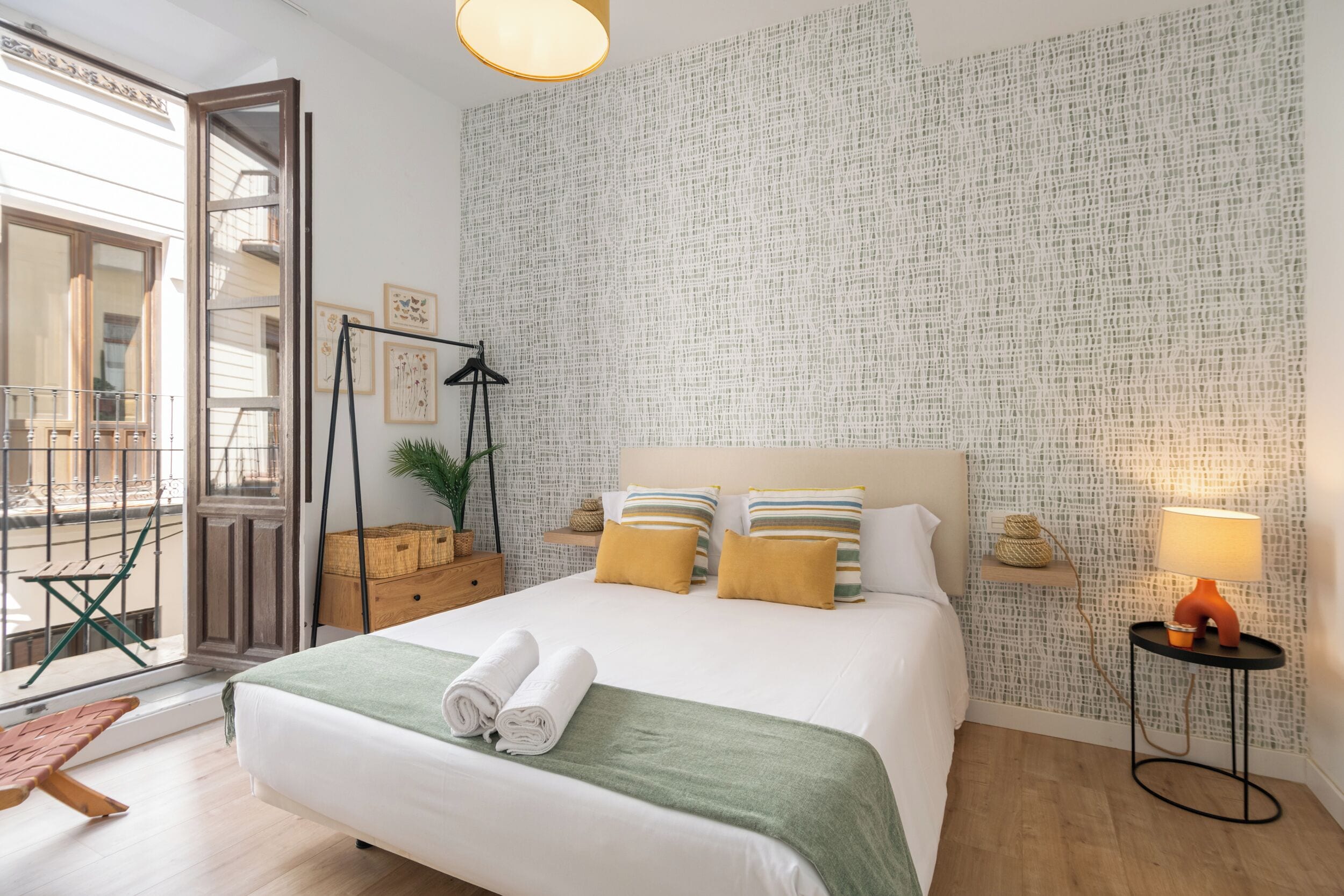 A neatly arranged bedroom with a large bed, a side table, a hanging plant, and a view through an open door.