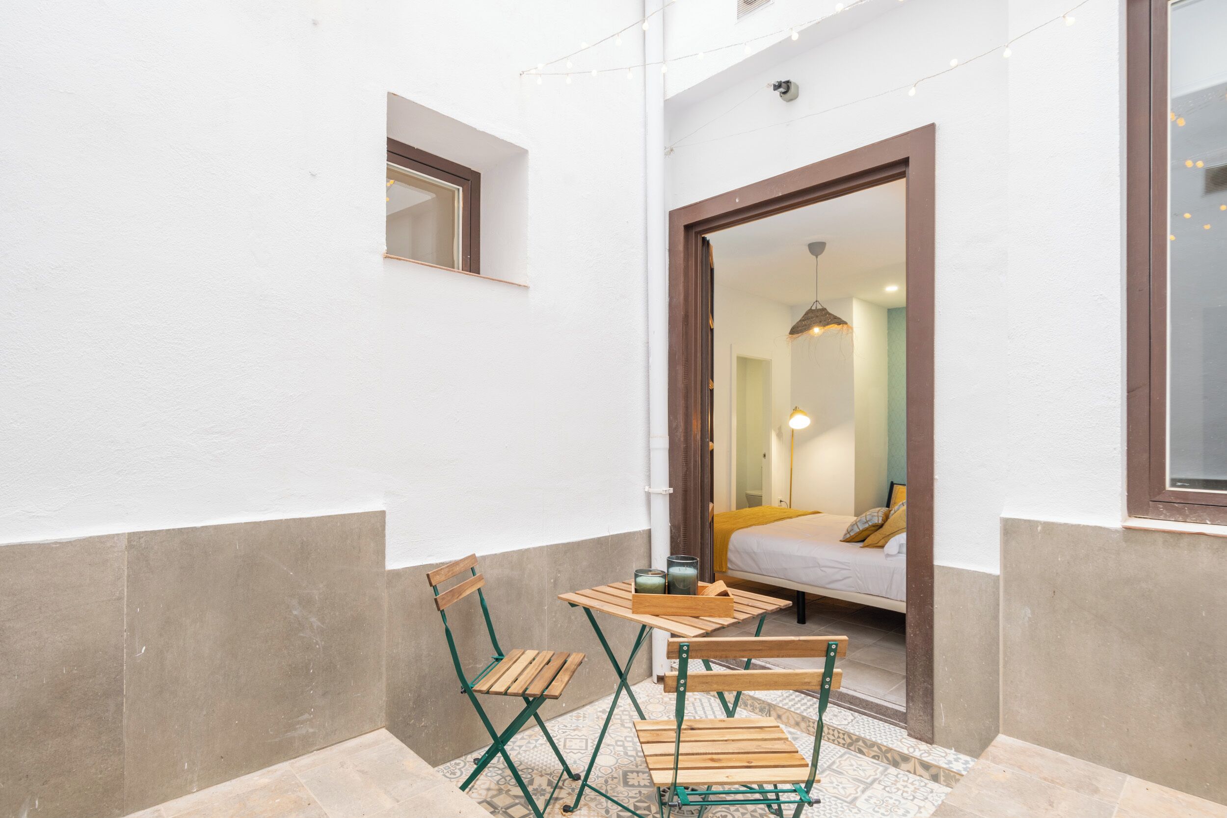 A small outdoor seating area with a table and two chairs, a doorway leading to a bedroom, and a string of lights above.
