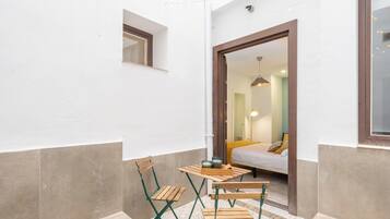 Un pequeño espacio al aire libre con una mesa y dos sillas, un acceso a un dormitorio, y una guirnalda de luces colgando.