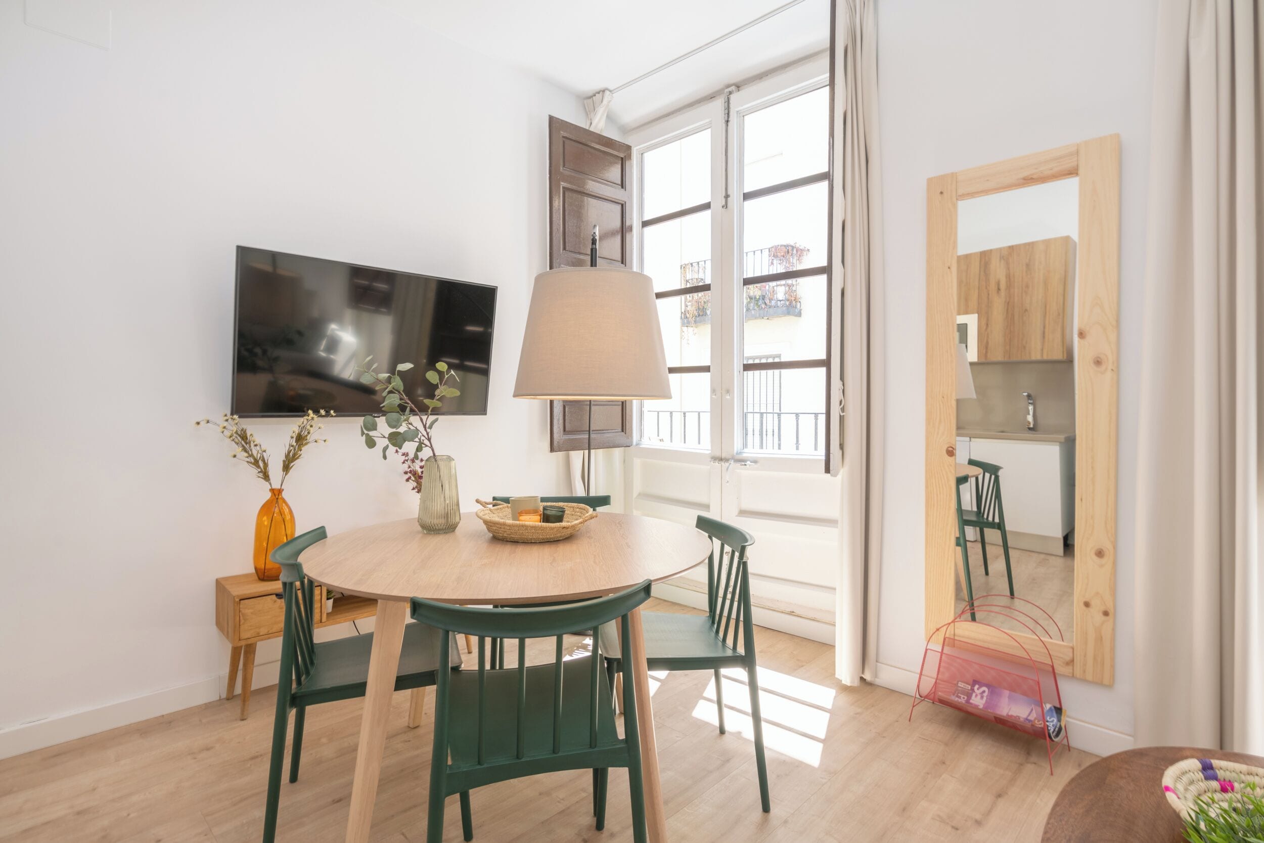 A dining area with a round table, chairs, a TV mounted on the wall, and a window with a view of balconies.