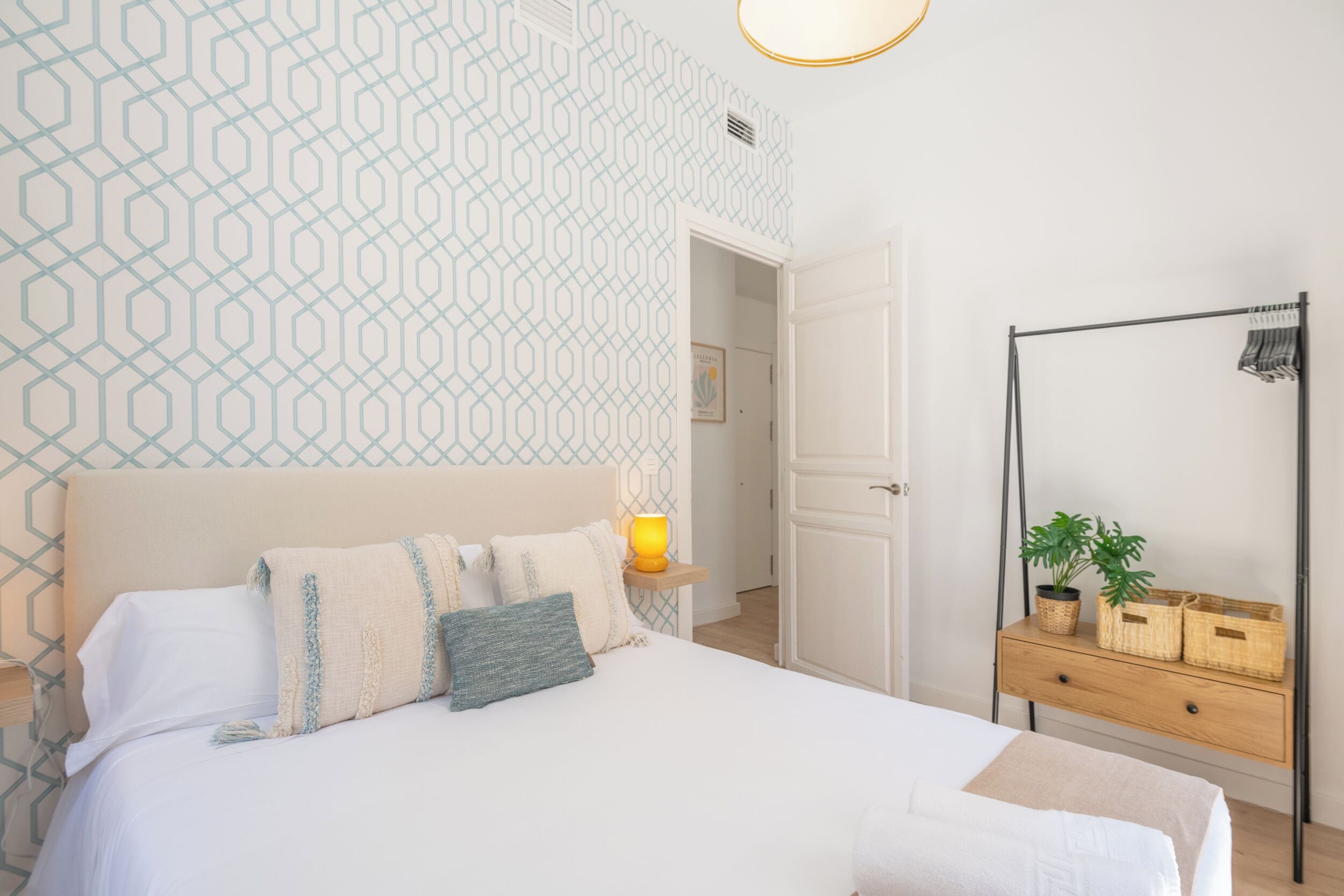 Une chambre à coucher bien rangée, avec un mur décoré d’un motif, une table de chevet avec une lampe, un miroir et une plante posée sur une commode.