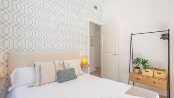 Une chambre à coucher bien rangée, avec un mur décoré d’un motif, une table de chevet avec une lampe, un miroir et une plante posée sur une commode.
