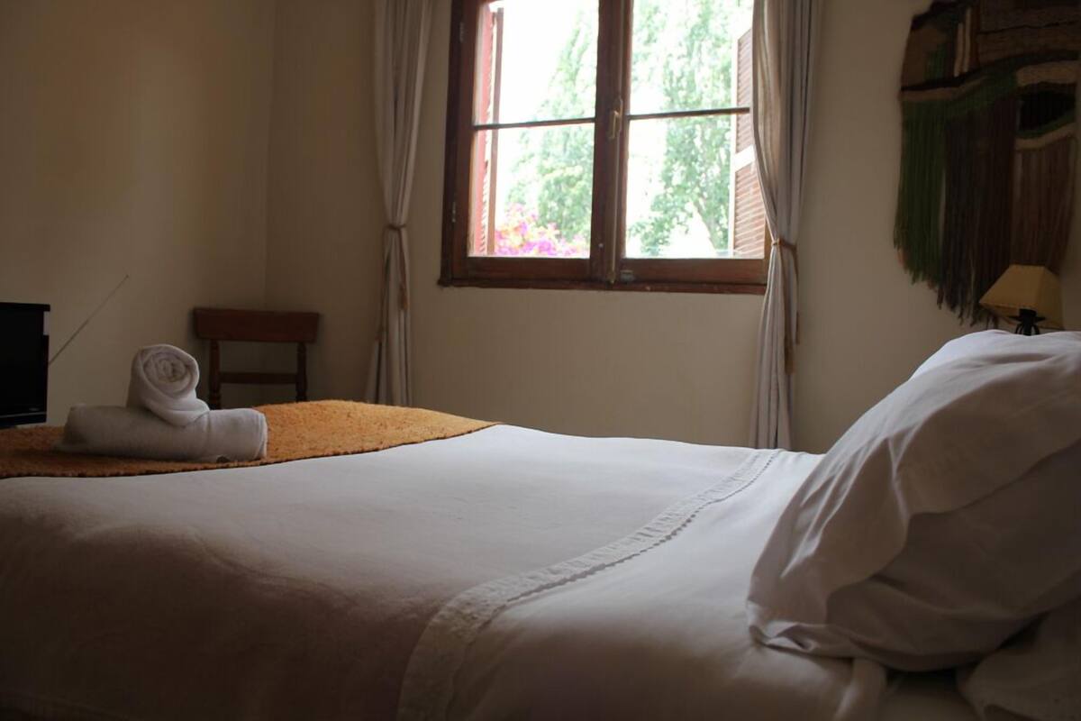 Una cama bien hecha con cubrecama y almohadas blancas, una toalla enrollada a un lado, una pequeña silla de madera y una ventana con cortinas.