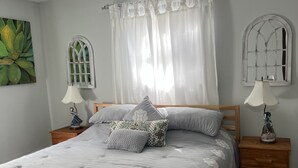 Una cama bien hecha con un edredón floral gris, dos mesitas de noche con lámparas, un ventanal con marco decorativo y un cuadro en la pared.