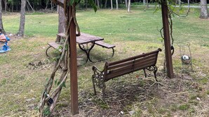 Una pérgola de madera con plantas trepadoras y flores rojas, un banco de madera y una mesa de picnic en un espacio al aire libre cubierto de césped.