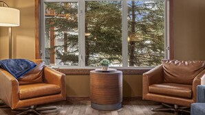 Een kamer met twee leren fauteuils, een klein houten bijzettafeltje met een plant, en een groot raam met uitzicht op bomen en gebouwen.