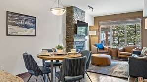 Een moderne woonkamer met een stenen openhaard, een eethoek met een ronde tafel en stoelen, en een groot raam met uitzicht op bomen.