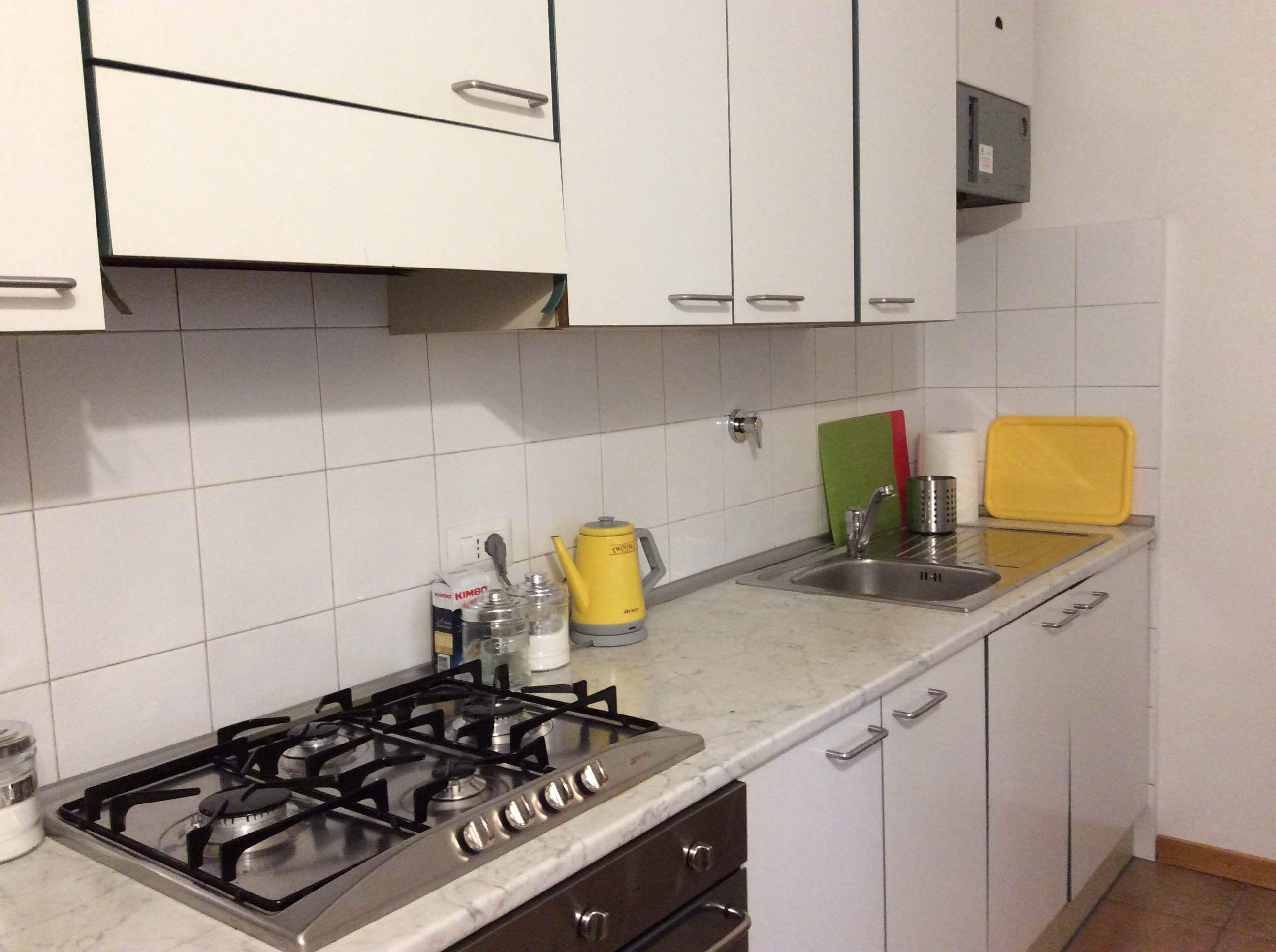Une cuisine avec des meubles blancs, une cuisinière à gaz, un évier et un plan de travail sur lequel se trouvent divers objets.
