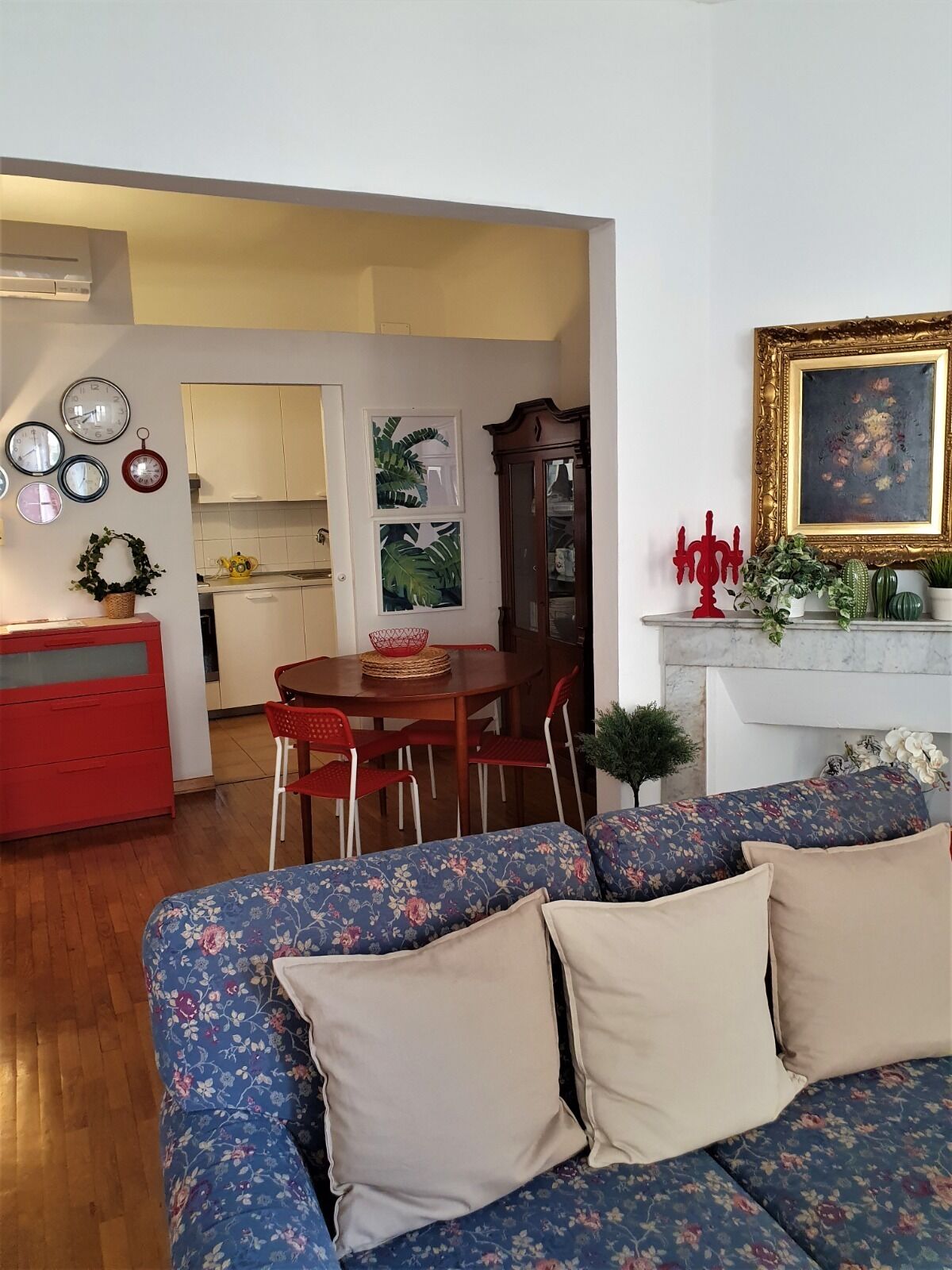 Un salon avec un canapé à motifs floraux, une table à manger avec des chaises, un coin cuisine et une cheminée agrémentée d’un tableau encadré.