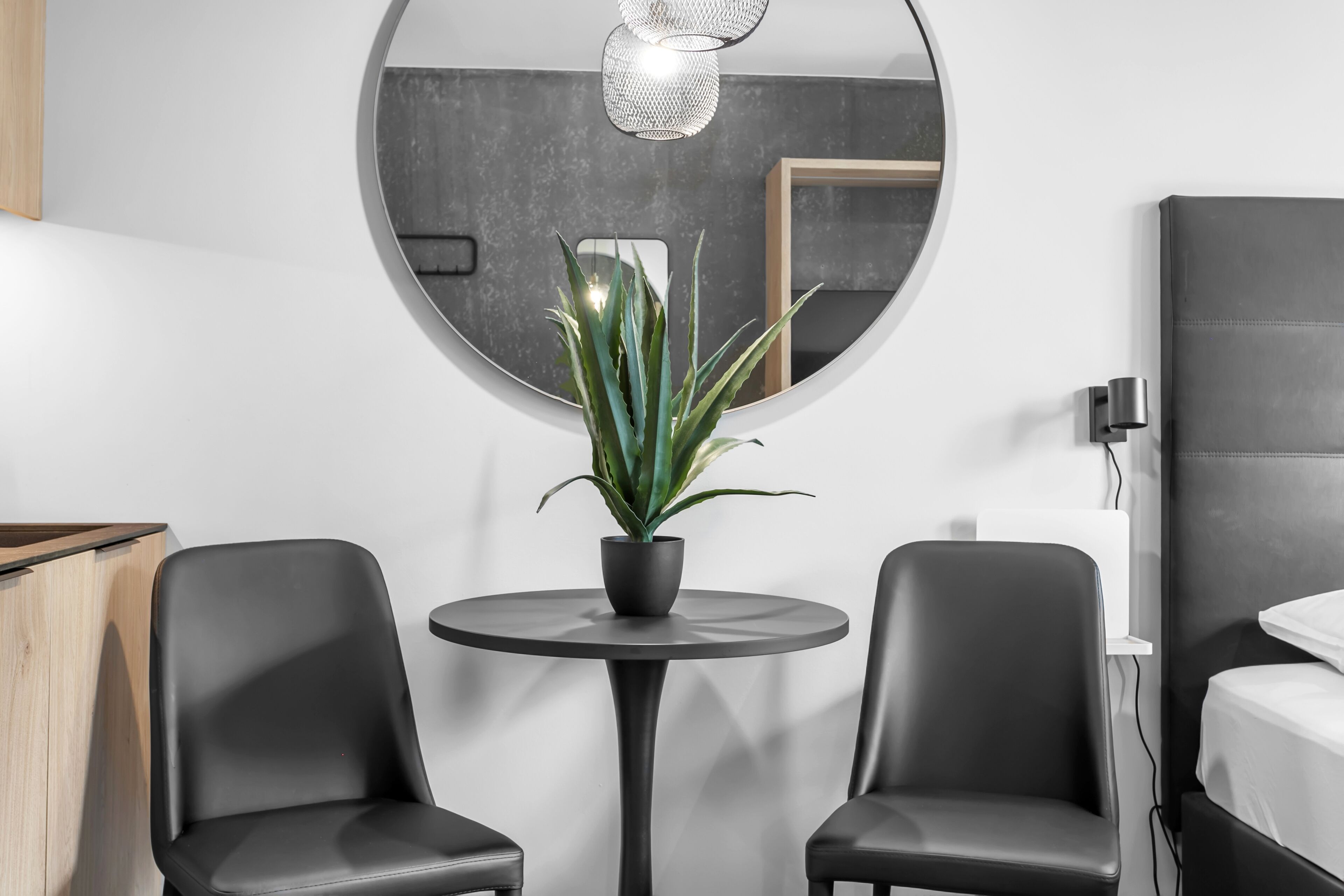 Une chambre moderne avec un grand miroir rond, deux chaises noires, une petite table sur laquelle est posée une plante en pot et un lit avec une tête de lit.