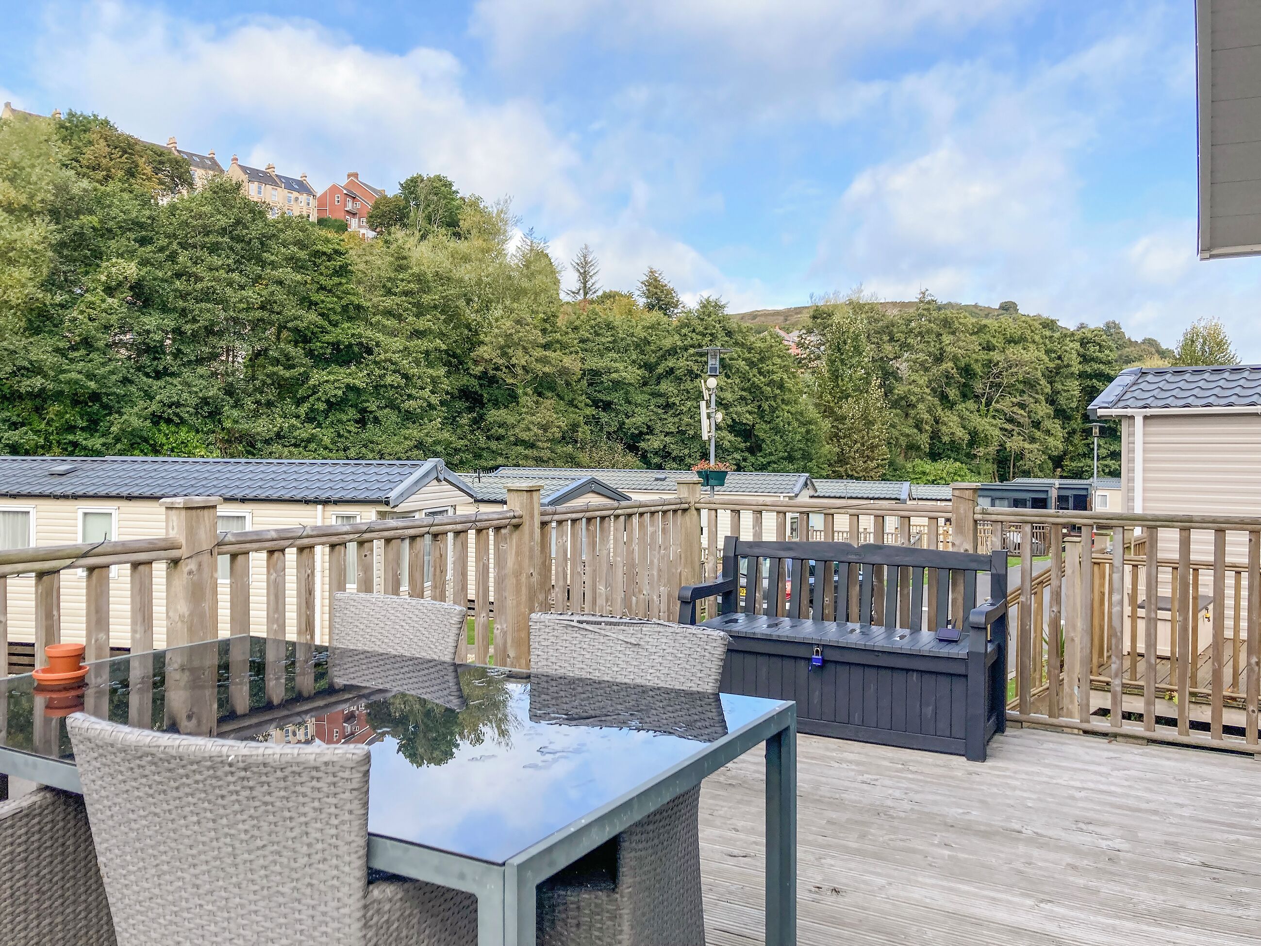 A wooden deck with a glass table and chairs, surrounded by a wooden fence, overlooking a forested area with buildings in the background.