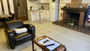 A modern living room with a leather sofa, a coffee table with remote controls and magazines, a kitchen with white cabinets and a microwave, and a fireplace with a brick surround.