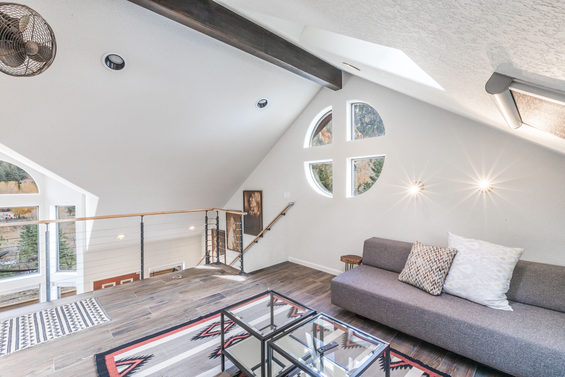 Um loft espaçoso com teto inclinado, uma mesa de centro de vidro, um sofá cinza com almofadas estampadas e vista para uma floresta através de amplas janelas.