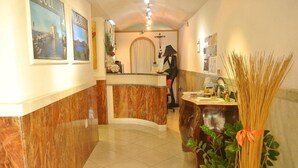 A hotel reception area with a marble reception desk, framed pictures on the wall, a cross, and a plant with a ribbon. - Hotel Vistamare (Pozzuoli)