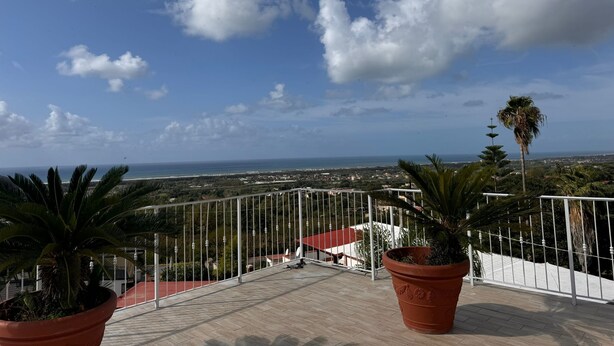 A terrace with potted plants, a white railing, and a view of the sea and distant buildings. - Hotel Vistamare (Pozzuoli)