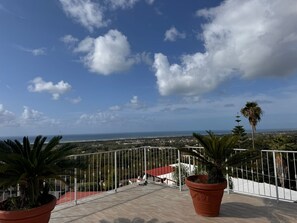 A terrace with potted plants, a white railing, and a view of the sea and distant buildings. - Hotel Vistamare (Pozzuoli)