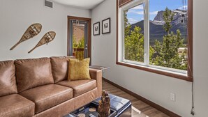 A living room with a brown leather sofa, a coffee table, and a large window with a mountain view. - 180° Mountain Views | Two Hot Tubs | New Design (Canmore)