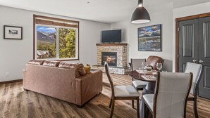 A living room with a stone fireplace, a sofa, a dining table with chairs, a TV mounted on the wall, and a large window with a mountain view. - 180° Mountain Views | Two Hot Tubs | New Design (Canmore)