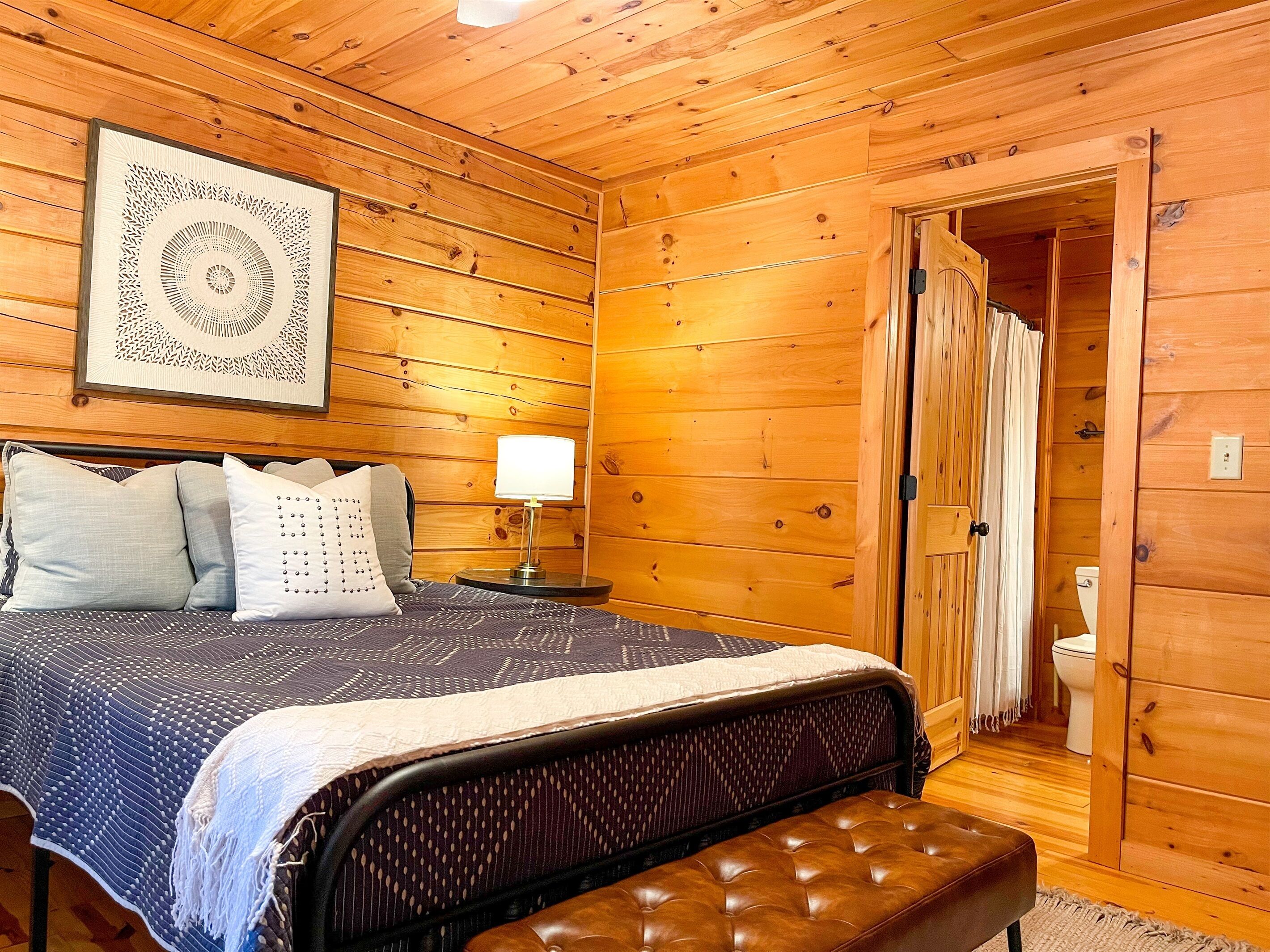 A wooden cabin bedroom with a bed, pillows, a bedside lamp, a bench, and a door leading to a bathroom.