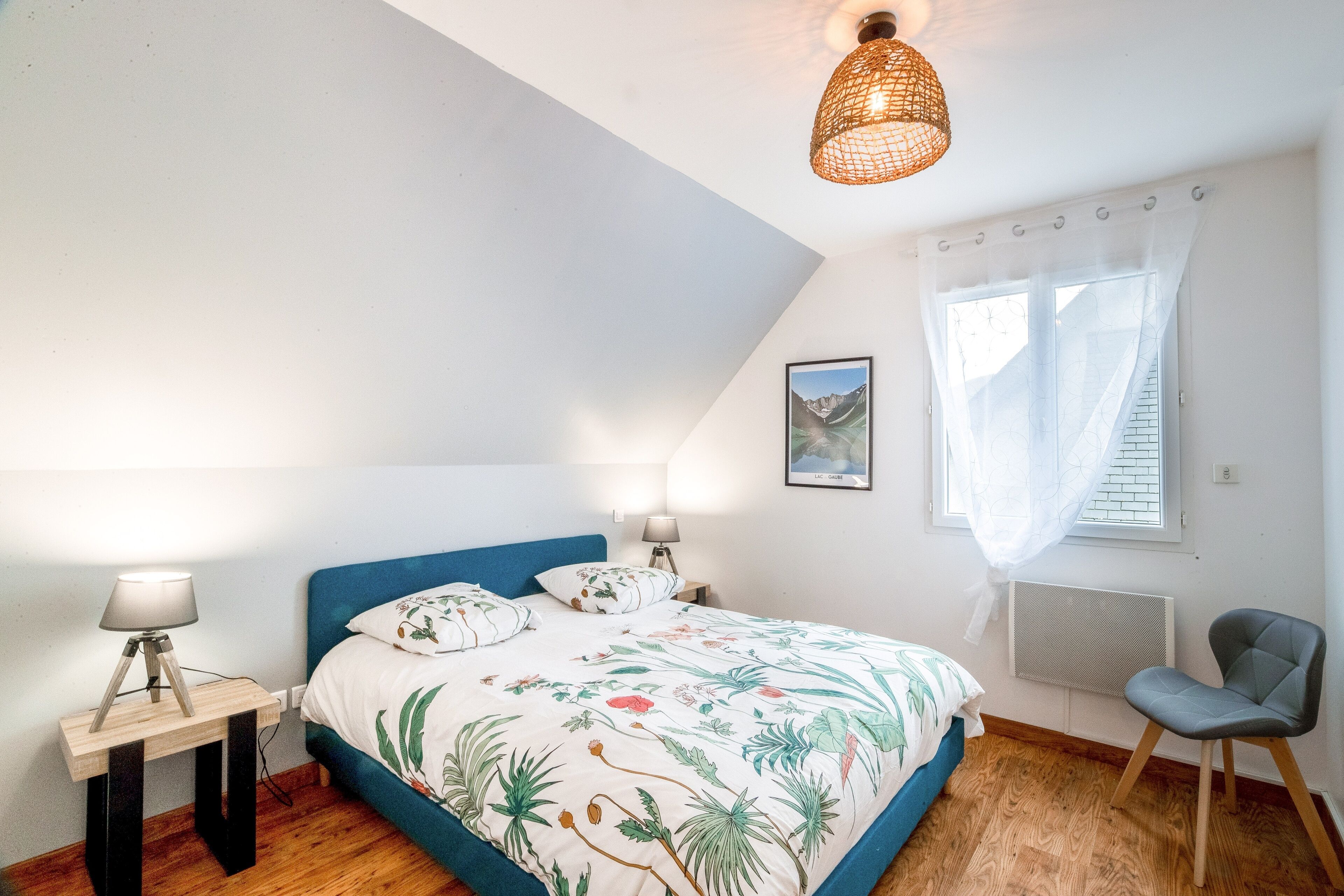 Een slaapkamer met een bed, een stoel, een nachtkastje, een lamp, een foto aan de muur en een raam met gordijnen.