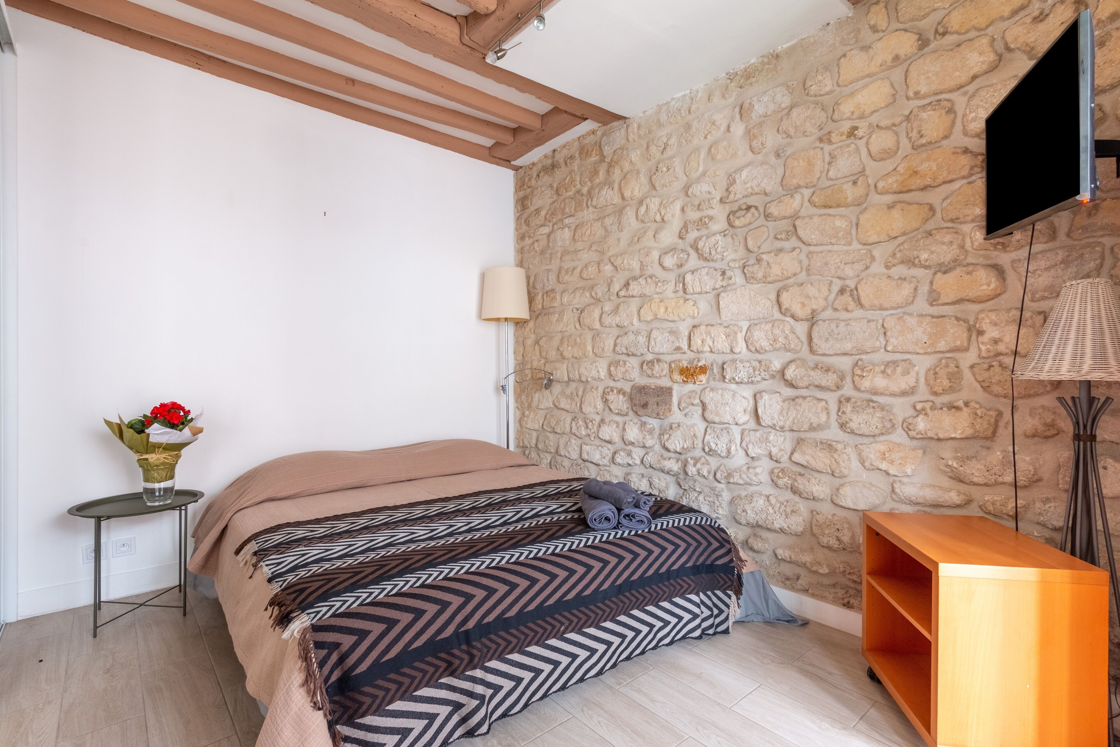 Une chambre avec un mur en pierre, un lit avec une literie à motifs, une petite table sur laquelle est posé un vase de fleurs, et un téléviseur fixé au mur.