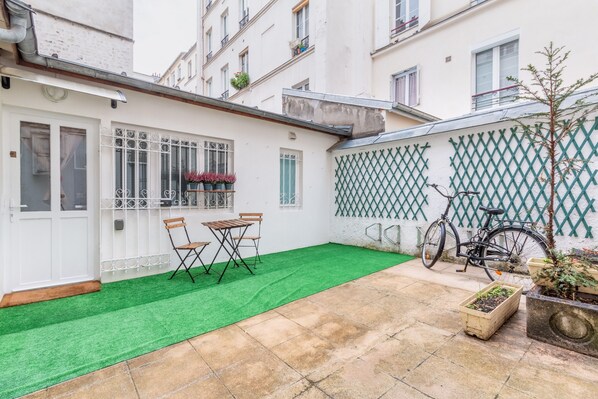 Un patio con césped artificial verde, una mesa y sillas de madera, una bicicleta y plantas en macetas.