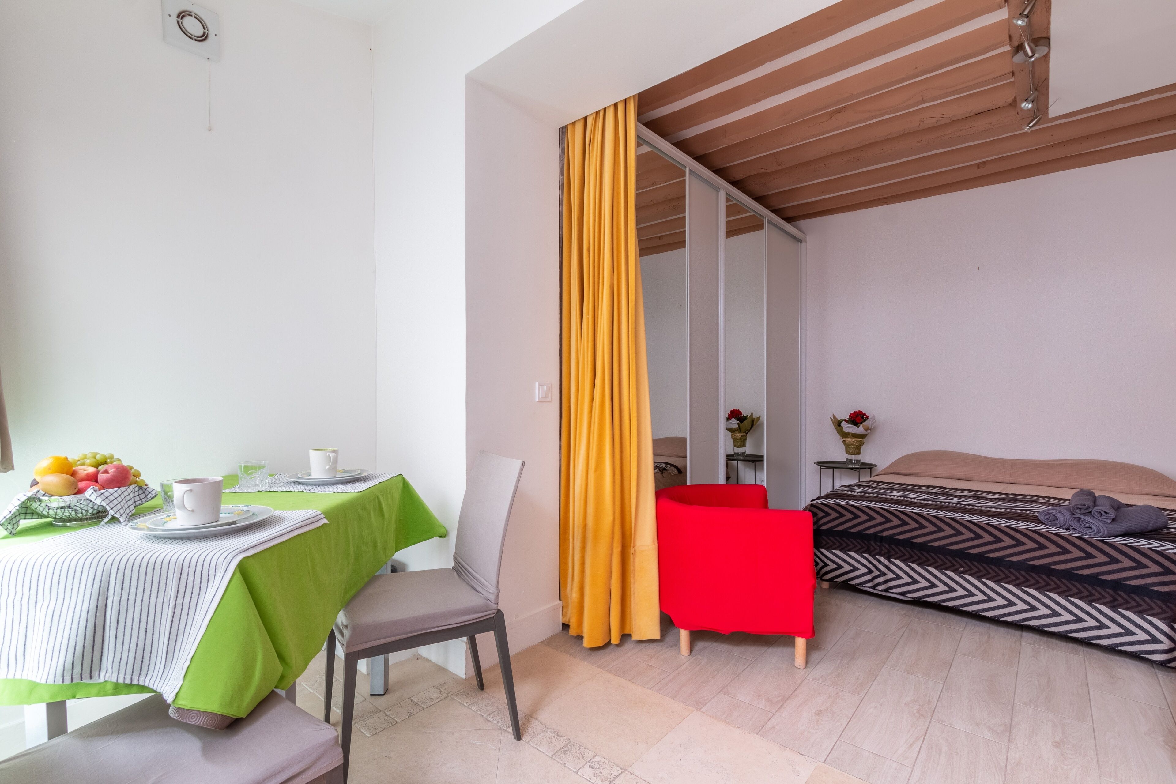 Une pièce avec une table à manger dressée pour deux personnes, un lit recouvert d’une courtepointe à motifs, une chaise rouge et un rideau jaune.
