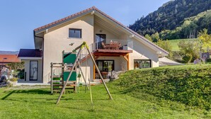 Ein zweistöckiges Haus mit Balkon, Spielplatz und Blick auf einen bewaldeten Hügel.