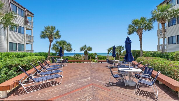 A wooden deck with lounge chairs and umbrellas, overlooking a beach with palm trees and a clear blue sky. - Ocean Forest Villas 115C- 2 Br Condo in Myrtle Beach (Myrtle Beach)