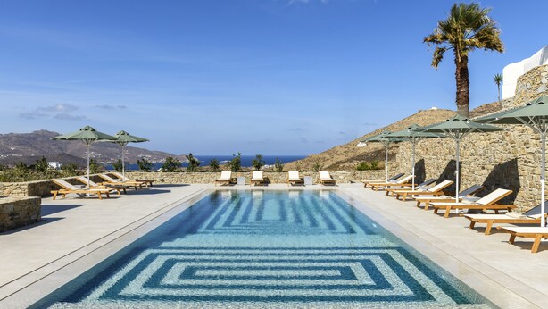 Una piscina con un diseño geométrico, rodeada de sillones de descanso y sombrillas, con vista al mar y a las montañas al fondo.