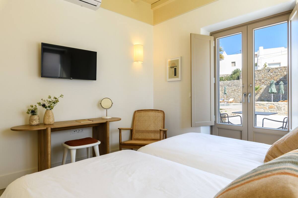 A hotel room with two beds, a TV, a desk with a chair, a small stool, and a window with a view of the outside.