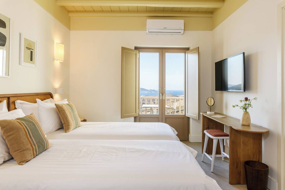 A hotel room with two beds, a desk, a TV, and a view of the city.