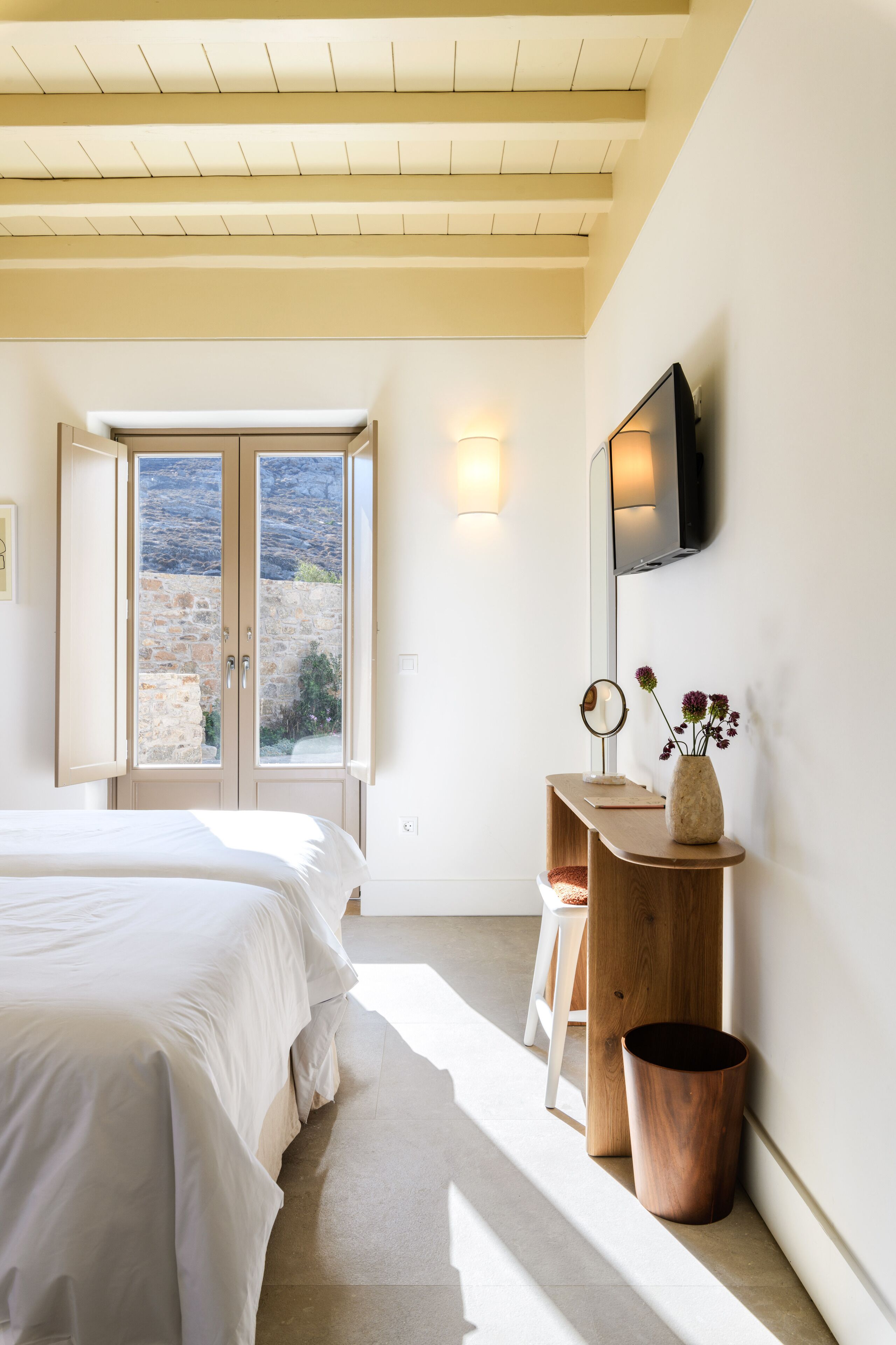 Une chambre à coucher avec un lit, un bureau en bois, un téléviseur fixé au mur et une fenêtre donnant sur un mur en pierre.