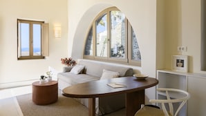 Una sala moderna con una mesa redonda de madera, un sofá blanco, una mesita auxiliar redonda de madera, una silla blanca, un ventanal arqueado grande y un aparador pequeño blanco con un marco de foto.