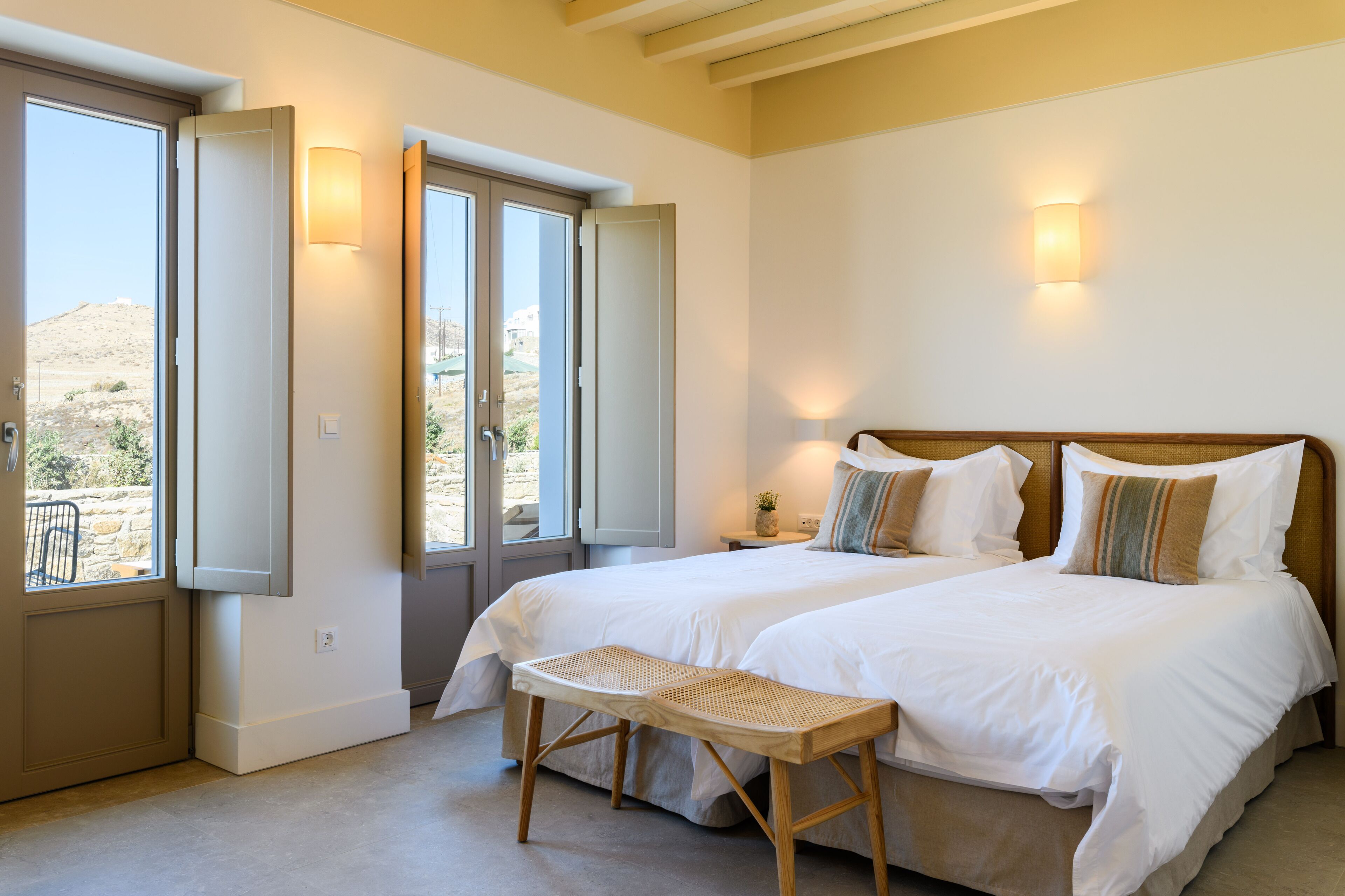 Un dormitorio con dos camas, un banco y vista a un paisaje desértico a través de puertas abiertas.