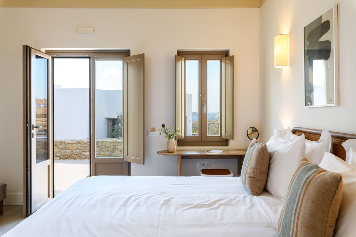 A bedroom with a bed, a bedside table, a dresser, a mirror, and a window with an open door leading to a balcony.