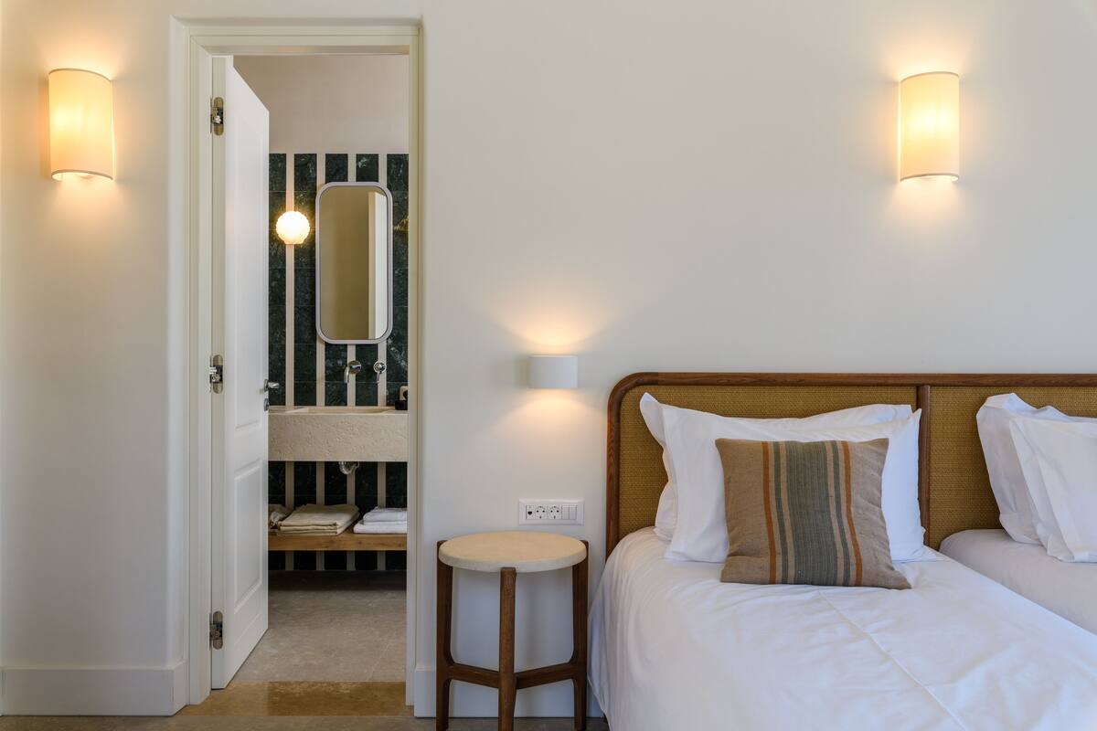 A hotel room with a bed, a round stool, a mirror, and two wall-mounted lights.