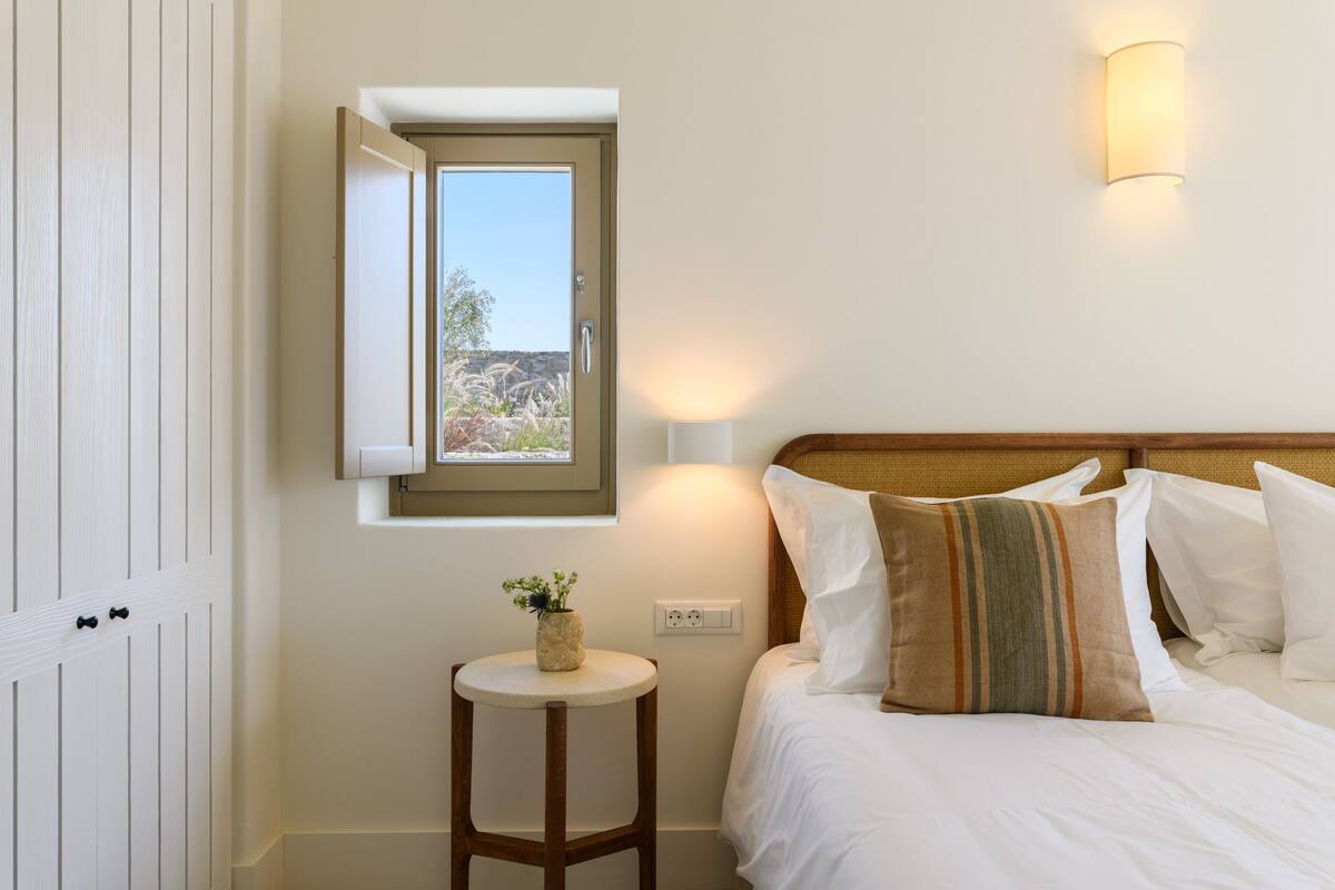 A bedroom with a bed, a bedside table with a vase, a window, and a wall-mounted light.